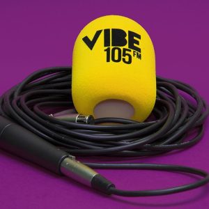 Yellow cylindrical foam microphone windscreen printed “VIBE 105 FM”, shown with a handheld microphone and coiled cable on a purple background.
